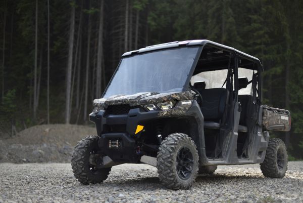 Can-Am Defender side-by-side utility vehicle with camouflage body parked on rocky ground in front of a forest.